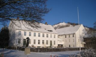 Bildet viser Andorsengården, Mandal museum, en fin vinterdag med små på taket og på bakken. I bakgrunnen ses utsiktspunktet Uranienborg.