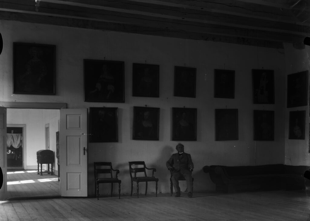 Adolph Arenfeldt (1824-1909) på Sæbygård. Han sitter i en sal med malerier på veggene.