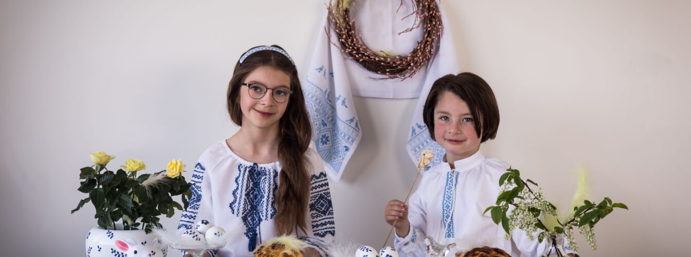 To jenter sitter ved et påskepyntet bord, kledd i tradisjonelle, broderte skjorter fra Ukraina
