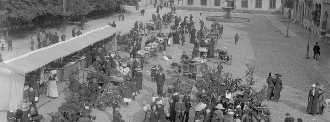 Bildet er i svart-hvitt og viser et mylder av mennesker som handler og prater sammen på torvet. Det er folk i alle aldere der. Ut fra klær og stil er bildet tatt cirka 1915.