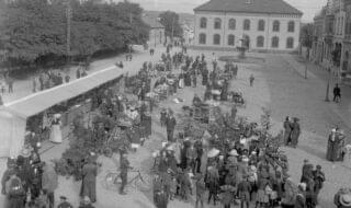 Bildet er i svart-hvitt og viser et mylder av mennesker som handler og prater sammen på torvet. Det er folk i alle aldere der. Ut fra klær og stil er bildet tatt cirka 1915.