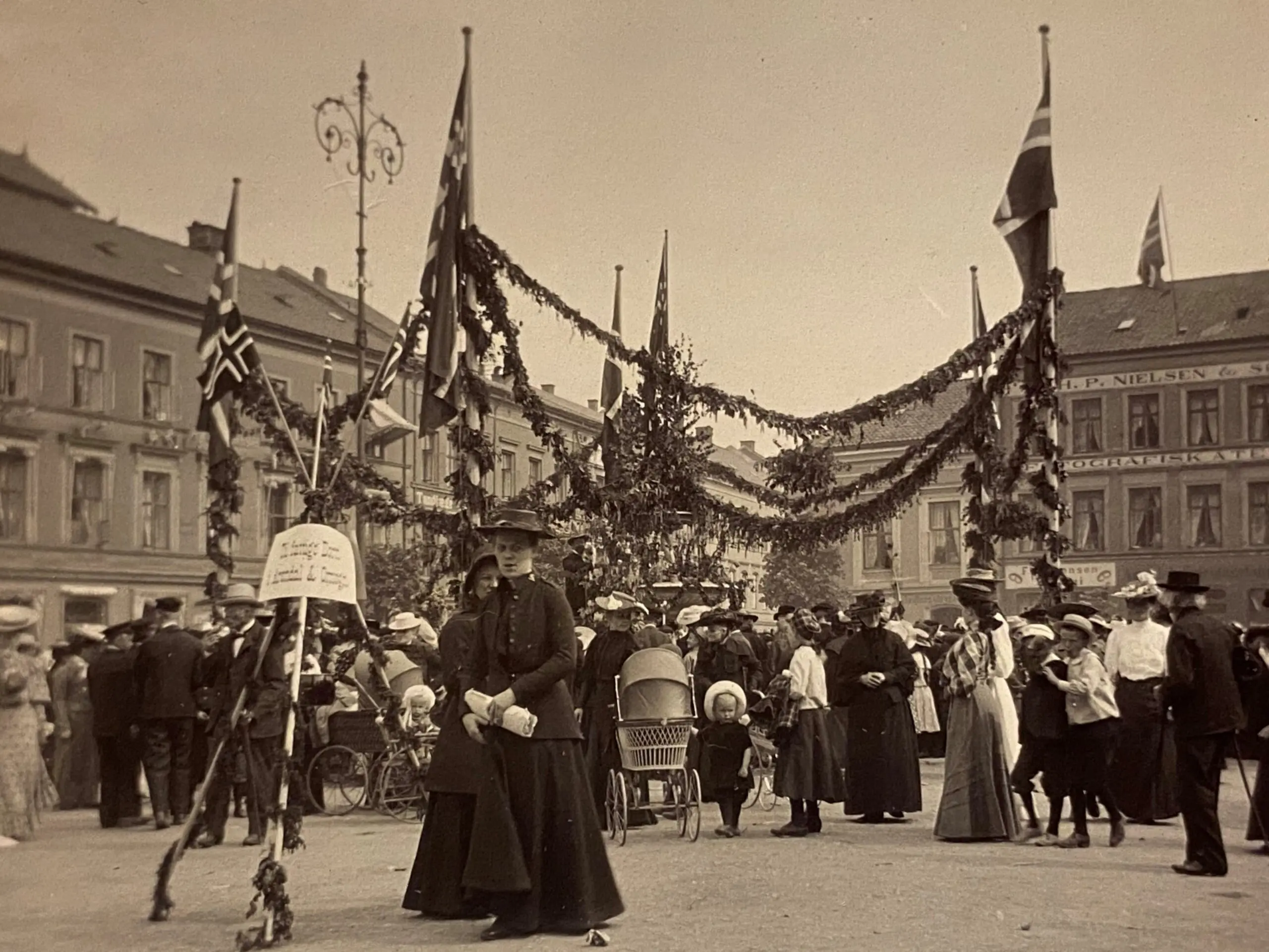 Sepiafarget foto fra 1905 som viser mennesker med norske flagg, girlandere av granbar under markering av Barnehjelpsdagen i Arendal. Alle bærer hatt, også barna. Kvinnene har lange kjoler. De er på en åpen plass.