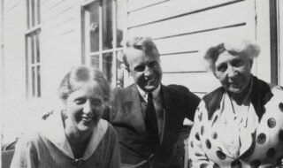 Svart-hvitt fotografi av tre mennesker sittende utenfor hvitt trehus med liggende panel. Fra venstre en ung kvinne, i midten en ung mann, en eldre kvinne til høyre.