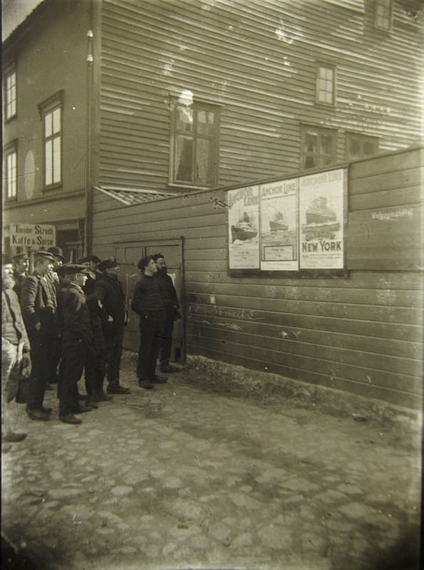 En gjeng skuelystne unge menn i Farsund ca. 1900 som kikker lengselsfullt på reklameplakater for Amerikabåten.