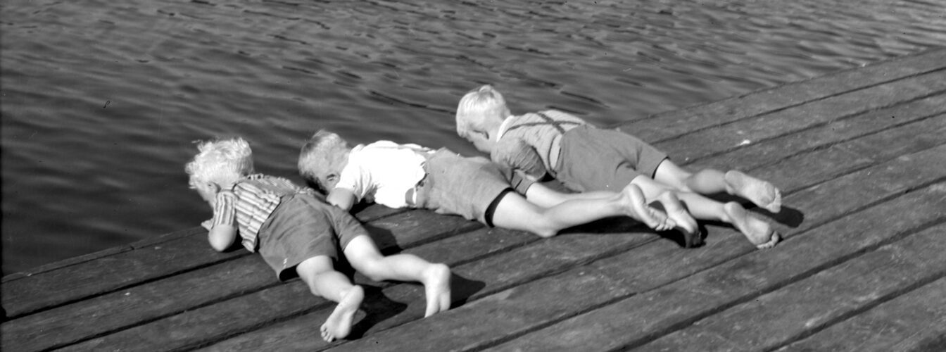 Svart/hvitt foto av barn på brygge som kikker ned i sjøen.